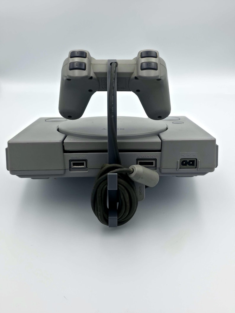 May include: A gray PlayStation console with a matching controller. The controller is connected to the console with a black cable. The console has a disc tray and several ports. The controller has a classic design with directional buttons and four action buttons.