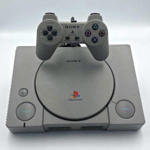 May include: A gray PlayStation console with a matching controller. The console has the PlayStation logo and the word "SONY" printed on it. The controller is placed on top of the console.