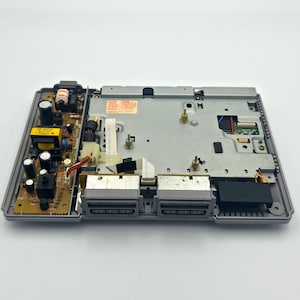 May include: An open, gray PlayStation 1 console, revealing internal components. The console's interior includes a circuit board with various electronic parts, connectors, and a power supply. Two controller ports are visible on the front.