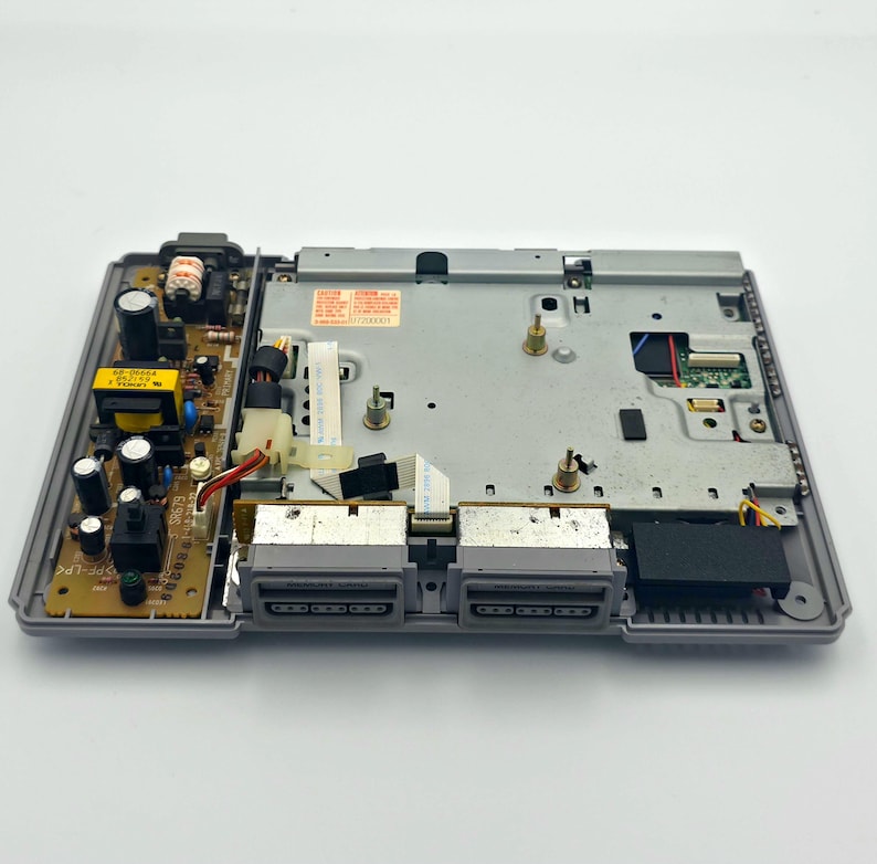 May include: An open, gray PlayStation 1 console, revealing internal components. The console's interior includes a circuit board with various electronic parts, connectors, and a power supply. Two controller ports are visible on the front.