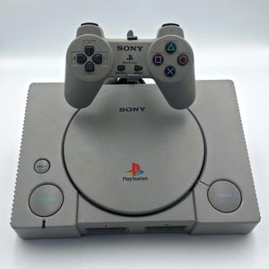 Könnte beinhalten: Eine graue Sony PlayStation-Konsole mit passendem Controller. Die Konsole hat ein Laufwerk, Ein- und Aus-Tasten sowie das PlayStation-Logo. Der Controller hat Richtungstasten, Aktionstasten und das Sony-Logo.