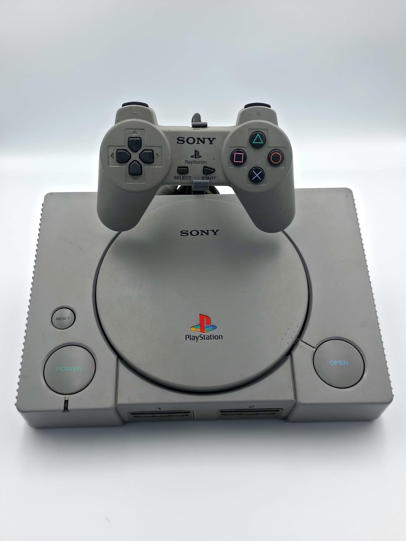 May include: A gray Sony PlayStation console with a matching controller. The console has a disc tray, power and reset buttons, and the PlayStation logo. The controller has directional buttons, action buttons, and the Sony logo.