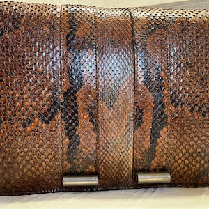 Peut inclure: Une pochette rectangulaire en peau de serpent marron et noire avec une fermeture à rabat et une quincaillerie argentée. Le sac présente un motif texturé et écailleux et une longue et fine bandoulière. Accessoire vintage.