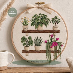 Op de afbeelding: Een ronde houten borduurring met een stoffen paneel met handgestikte ontwerpen van zes potplanten op twee bruine planken. De bovenste plank bevat madeliefjes, een groene plant met bladeren en een kleine boom. De onderste plank toont een monstera, een vetplant en een hangplant met roze bloemen. Bovenaan is een beige lint gestrikt.