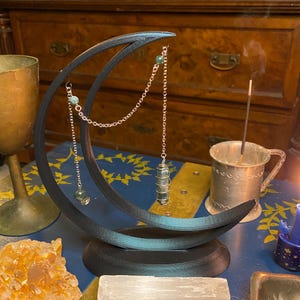 May include: A black crescent moon-shaped stand holding a silver chain with a green crystal pendant and small green beads. The stand is positioned on a blue surface alongside a large orange crystal cluster, a white selenite bar, and a metal cup.