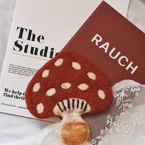 May include: A felt mushroom-shaped brooch with a red cap and white spots. The stem is beige with black and white gills. The brooch rests on a white lace surface, with a red book that says "RAUCH" and a magazine that says "The Studi" in the background.
