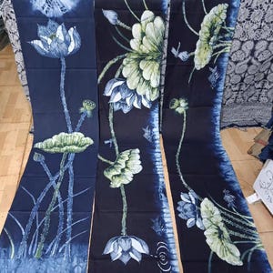 May include: Three dark blue fabric panels with a floral design. The panels feature white and blue lotus flowers, green leaves, and stems. The fabric has a tie-dye effect along the edges, creating a unique visual texture. The overall design is artistic and nature-inspired.