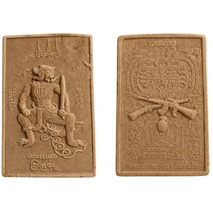 May include: Two rectangular, light brown amulets. One features a seated figure with a sword, surrounded by script. The other displays crossed rifles, a bottle, and script. Both have detailed relief carvings.