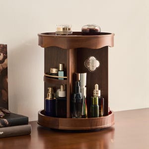 May include: A dark brown wooden rotating cosmetic organizer with multiple circular tiers and vertical dividers. The unit features a top shelf, a middle shelf with a gold-toned rail, and a base, holding various glass perfume and skincare bottles.