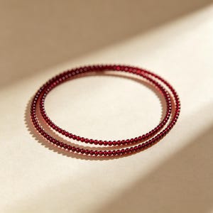 May include: A double-strand bracelet composed of small, uniform, deep red spherical beads. The beads have a smooth, polished finish and are strung together in two circular loops that overlap slightly, creating a consistent, minimalist design.