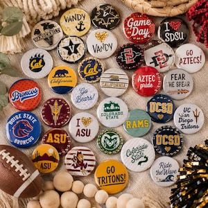 May include: A collection of circular pin-back buttons featuring various university and sports team logos, including Vanderbilt, SDSU, Cal, Boise State, ASU, Colorado State, and UCSD, with some designs accented by small metallic studs or heart shapes.
