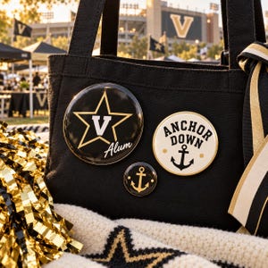 May include: A black canvas tote bag adorned with three circular pin-back buttons. One large black button features a gold star with a white V and the text Alum. A cream button displays the text Anchor Down with an anchor icon. A small black button features a gold anchor.