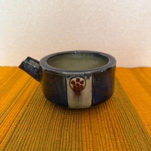 Puede incluir: Un pequeño cuenco de cerámica hecho a mano con un esmalte azul oscuro. Cuenta con un pico y una franja vertical decorativa con un diseño de huella de pata. El cuenco está sobre una superficie amarilla texturizada.