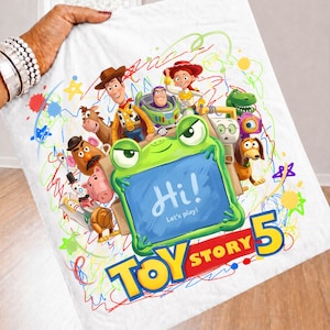 May include: A white fabric item featuring a colorful Toy Story 5 graphic. The design includes characters like Woody, Buzz Lightyear, and Jessie surrounding a green frog-shaped frame with the text Hi! Let's play! and Toy Story 5 below.