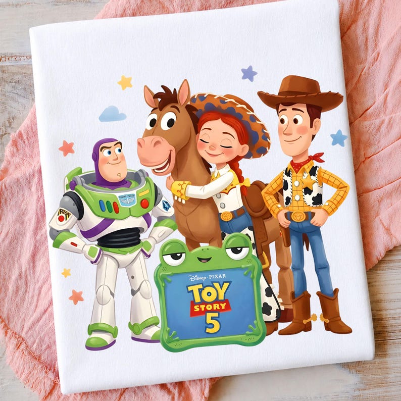 May include: A white t-shirt featuring a colorful graphic of Buzz Lightyear, Jessie, Bullseye, and Woody from Toy Story. A green frog-shaped frame in the foreground displays the text Disney Pixar TOY STORY 5, surrounded by small stars and clouds.