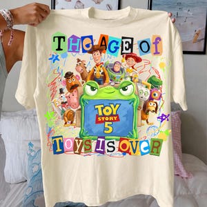 May include: A cream-colored t-shirt featuring a colorful graphic print of Toy Story characters including Woody, Buzz Lightyear, and Jessie. The design includes a green frog frame, scribbled lines, and the text "THE AGE OF TOY STORY 5 TOYS IS OVER."