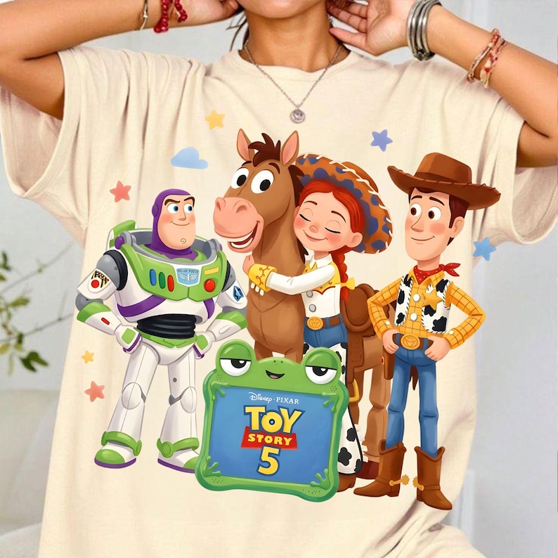 May include: A cream-colored t-shirt featuring a graphic print of Buzz Lightyear, Bullseye, Jessie, and Woody from Toy Story. A green frog-shaped frame in the foreground displays the text Disney Pixar Toy Story 5, surrounded by small stars.