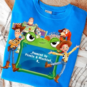 May include: A bright blue t-shirt featuring a graphic print of Woody, Buzz Lightyear, Jessie, and Slinky Dog from Toy Story surrounding a green frog-shaped frame. The frame contains the text _Powered by Pixels & Mischief_.