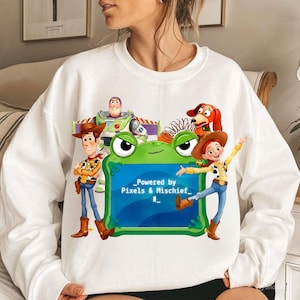 May include: A white long-sleeve sweatshirt featuring a graphic of Woody, Buzz Lightyear, Slinky Dog, and Jessie from Toy Story surrounding a green frog-shaped frame with the text "Powered by Pixels & Mischief" printed in the center.