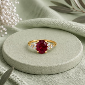 May include: A gold ring featuring a large oval-cut deep red gemstone at the center, flanked by three small round clear stones on each side. The band is thin and polished gold, showcasing a classic prong setting for the central red stone.