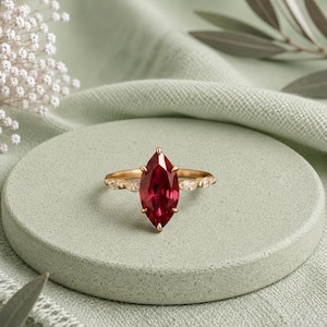 Puede incluir: Un anillo de oro con una gran piedra preciosa de color rojo intenso en talla marquesa como pieza central, flanqueada por dos piedras claras más pequeñas en talla marquesa a cada lado de la banda, con un engaste que resalta el color.