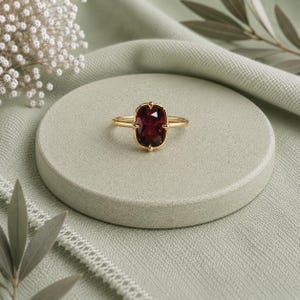 May include: A gold ring featuring a deep red, cushion-cut gemstone set in a four-prong mounting. The band is thin and polished, showcasing a classic design that highlights the vibrant color and faceted surface of the central stone.