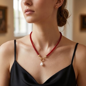 Op de afbeelding: Een ketting met een streng kleine, ronde, rode kralen en een goudkleurige knevelsluiting in het midden. Een enkele, witte, druppelvormige parel hangt aan de knevelsluiting en vormt een blikvanger aan de rode kralenketting.