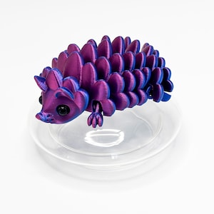 May include: A 3D printed articulated hedgehog figurine featuring a metallic iridescent finish that shifts between deep purple and vibrant blue. The creature has large black eyes and a segmented body with rounded, overlapping scales along its back.