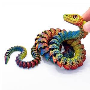 May include: A multi-coloured articulated snake toy featuring a gradient of red, yellow, green, and blue scales. The snake has a textured, segmented body design that allows for flexible movement, a detailed head with prominent eyes, and a tapered tail.