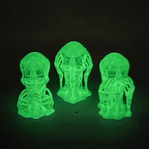 Puede incluir: Tres figuras que brillan en la oscuridad representando las poses de no ver, no oír y no hablar. Cada figura presenta una cabeza texturizada similar a un alienígena con rastas y un torso esquelético, emitiendo una luz verde neón brillante en la oscuridad.