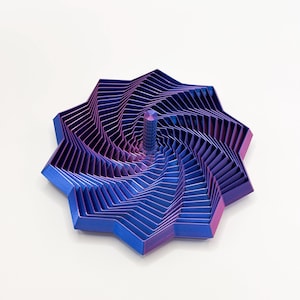 Puede incluir: Una escultura geométrica con un diseño en espiral. La pieza presenta un degradado de colores, que transita del azul al morado. Un elemento central, cilíndrico, se eleva desde el centro de la espiral. La escultura está sobre un fondo blanco.