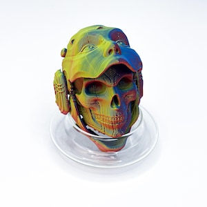 Puede incluir: Una escultura de cráneo impresa en 3D con un degradado de colores arcoíris que incluye amarillo, azul y rojo. El cráneo lleva un casco con accesorios laterales y descansa sobre una base de vidrio circular transparente.
