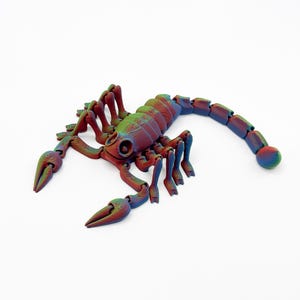 Peut inclure: Une figurine de scorpion articulée multicolore présentant un dégradé de teintes rouges, vertes et bleues. L'objet affiche un motif de surface texturé et superposé sur tout le corps, y compris les pinces, les multiples pattes et la queue segmentée.