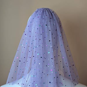 May include: A lilac tulle veil decorated with iridescent heart-shaped sequins. The veil is gathered at the crown and drapes down, creating a soft, romantic look. The heart-shaped sequins add a touch of sparkle and whimsy to the design.