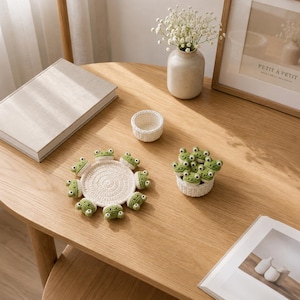 May include: A round, cream-colored crocheted coaster featuring eight small green frog heads attached to the perimeter, accompanied by a small, matching cream-colored crocheted bowl filled with several additional small green crocheted frog heads.