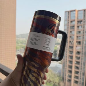 May include: A stainless steel travel mug with a black handle and lid. The mug features a tiger stripe design in shades of red, brown, and gold. A white label with text is wrapped around the mug.