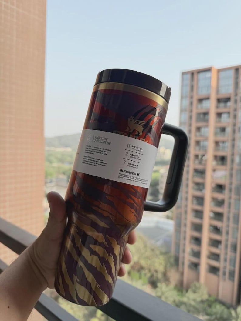 May include: A stainless steel travel mug with a black handle and lid. The mug features a tiger stripe design in shades of red, brown, and gold. A white label with text is wrapped around the mug.