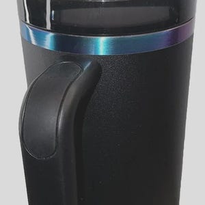May include: Black insulated tumbler with a handle and a clear lid. The tumbler has a black straw and a band of iridescent color around the middle. The bottom of the tumbler has a textured design.