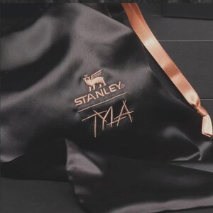 May include: Black satin bag with a copper-colored ribbon and embroidered logo. The logo features a winged animal above the word "STANLEY" and the initials "TMA" in a stylized font. The bag is likely for a Stanley product.