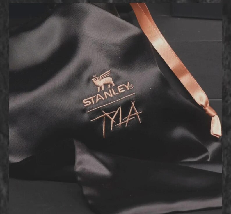 May include: Black satin bag with a copper-colored ribbon and embroidered logo. The logo features a winged animal above the word "STANLEY" and the initials "TMA" in a stylized font. The bag is likely for a Stanley product.