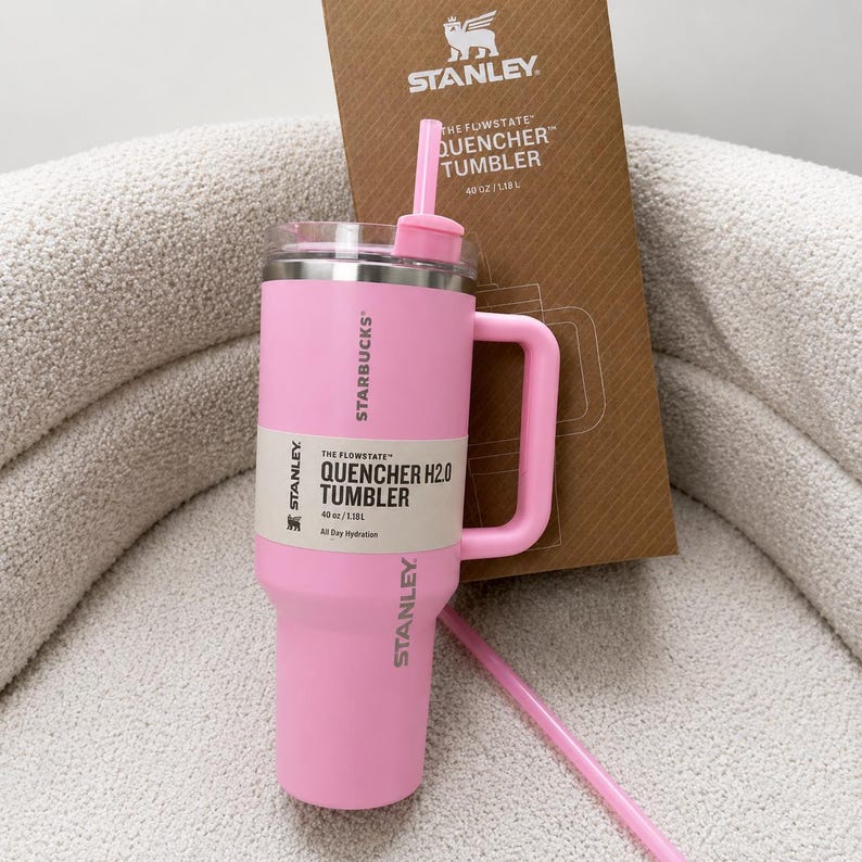 May include: A light pink Stanley Starbucks Flowstate Quencher H2.0 tumbler with a matching handle, lid, and straw. The tumbler features the Stanley and Starbucks logos, with a label stating 40 oz/1.18 L. A brown cardboard packaging box is behind it.