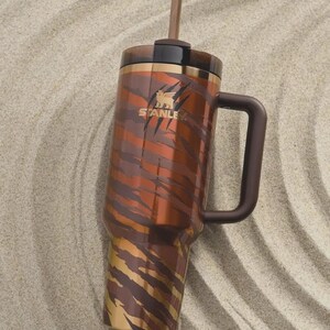 May include: A large, insulated tumbler with a handle and straw. The tumbler features a brown and tan tiger stripe pattern. The Stanley logo is visible on the side. The tumbler is designed to keep drinks cold or hot.
