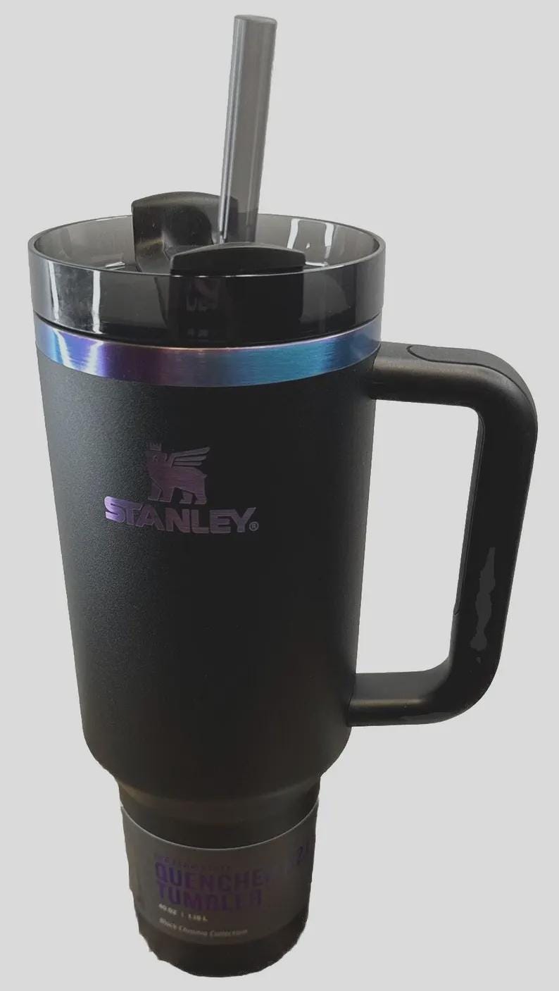 May include: Black Stanley Quencher tumbler with a handle and a clear straw. The tumbler has a black lid and a band of iridescent color around the middle. The Stanley logo is visible on the front.