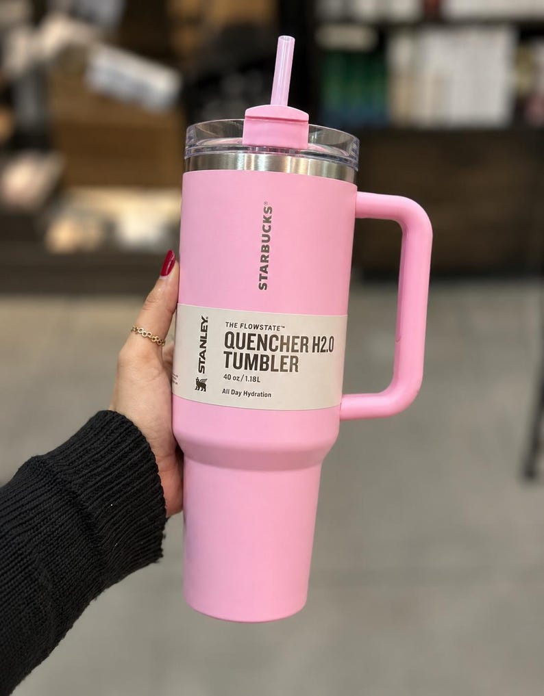 May include: A light pink Stanley Starbucks Quencher H2.0 tumbler with a 40 oz capacity. The cup features a matching pink handle, a clear lid with a pink straw, and a product label that reads The Flowstate Quencher H2.0 Tumbler 40 oz/1.18L.