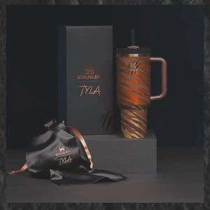 May include: A Stanley Tyla tumbler with a tiger stripe design in shades of brown and orange, a matching handle, and a black lid with a straw. The tumbler is displayed with a black gift box and a black satin bag, both featuring the Stanley logo.
