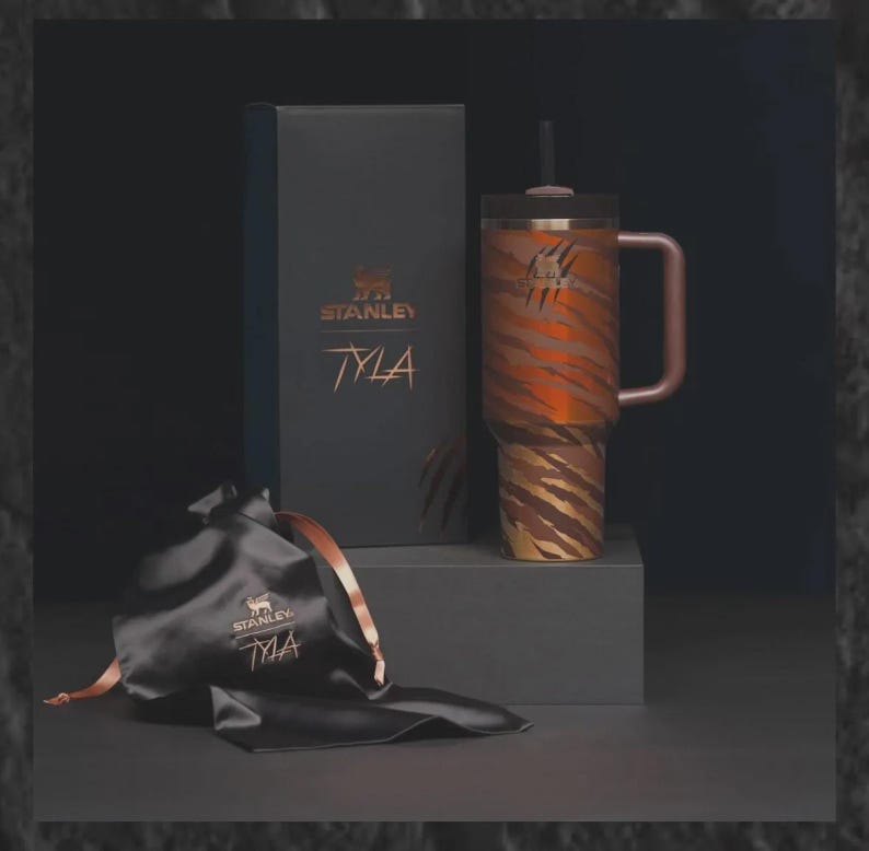 May include: A Stanley Tyla tumbler with a tiger stripe design in shades of brown and orange, a matching handle, and a black lid with a straw. The tumbler is displayed with a black gift box and a black satin bag, both featuring the Stanley logo.