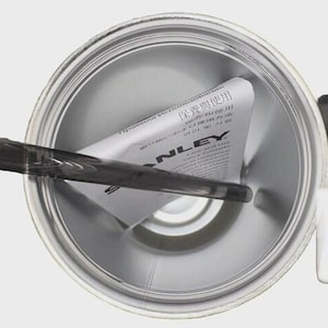 May include: Overhead view of a silver Stanley thermos. A black handle and a black and gray stirring stick are visible. A white label with Japanese text is inside the thermos.