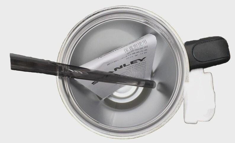 May include: Overhead view of a silver Stanley thermos. A black handle and a black and gray stirring stick are visible. A white label with Japanese text is inside the thermos.