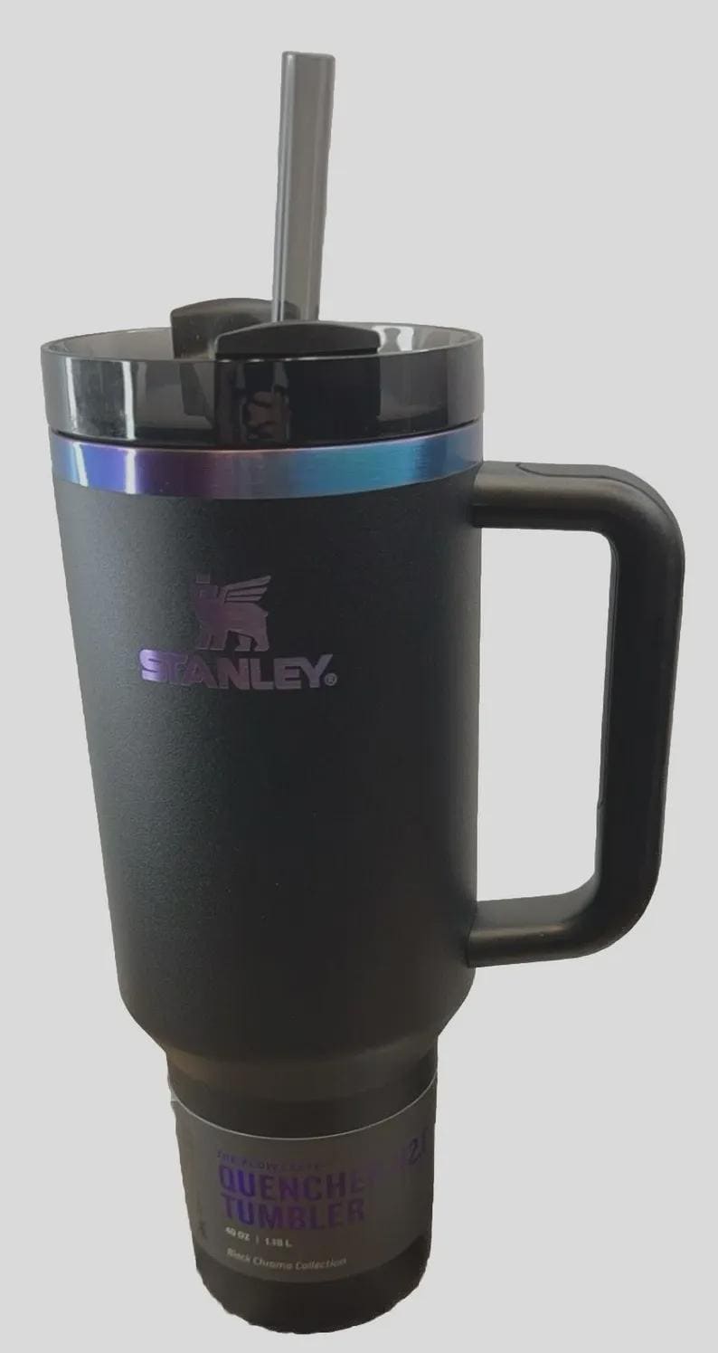 May include: Black Stanley Quencher Tumbler with a handle and straw. The tumbler has a black lid with a silver and blue iridescent band. The Stanley logo is visible on the front. The tumbler is part of the Black Chrome Collection.