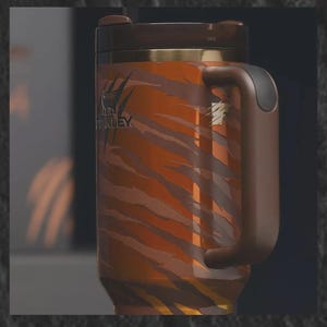 May include: A stainless steel travel mug with a tiger stripe pattern in shades of orange and brown. The mug has a brown handle, lid, and trim, and features the "Stanley" logo. The mug is designed for hot or cold beverages.